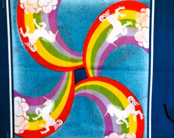 May include: A square, blue fabric with a white border featuring a whimsical design. The design includes four rainbow arcs, each with a white unicorn and fluffy clouds. The rainbows are red, orange, yellow, green, and purple.