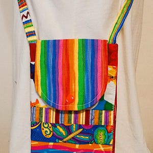 May include: A colorful crossbody bag with a rainbow striped flap and strap. The bag features a variety of patterned fabrics, including stripes, swirls, and cat motifs. The flap is secured with a red button. The strap is approximately 40 inches long.