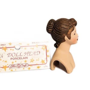 May include: A 3/4 inch porcelain doll head with brown hair in a bun. The doll head is on a white background with a packaging card that says "Doll Head Porcelain" and "Fibre-Craft".