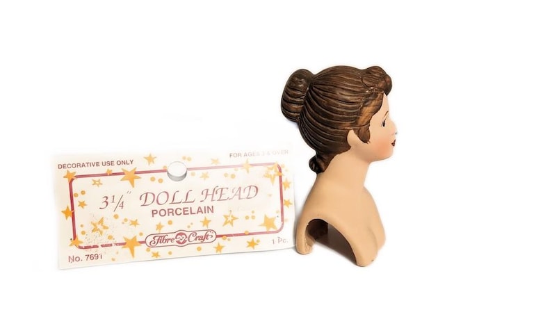 May include: A 3/4 inch porcelain doll head with brown hair in a bun. The doll head is on a white background with a packaging card that says "Doll Head Porcelain" and "Fibre-Craft".