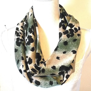 May include: An infinity scarf with a green, tan, and black animal print. The scarf is draped around a white mannequin bust. The fabric has a soft, flowing appearance, and the print features irregular black spots.