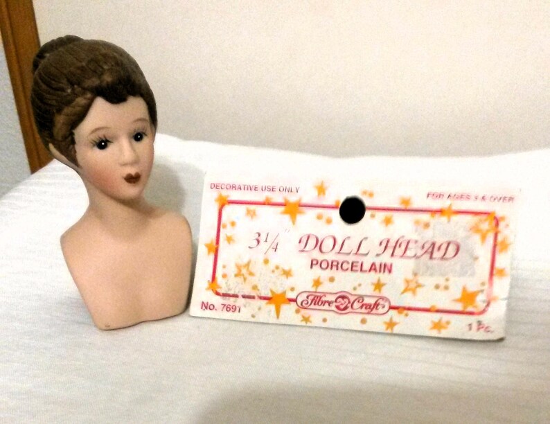 May include: A porcelain doll head with brown hair, a white bust, and a label that reads "3/4 Doll Head Porcelain Fibre-Craft No. 7691 1Pc".