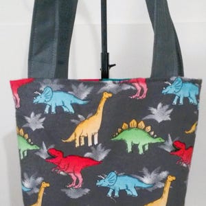 May include: A gray tote bag with a dinosaur print. The dinosaurs are in various colors, including red, blue, green, and yellow. The bag has gray straps.
