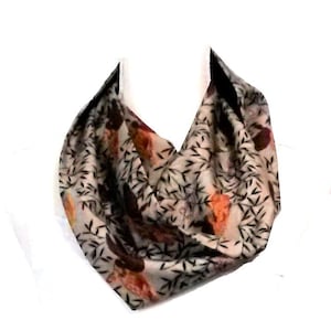 May include: A beige infinity scarf with a repeating pattern of orange, brown, and black botanical elements. The scarf is made of a soft, flowing fabric and is designed to be worn looped around the neck.