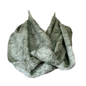 May include: A light green infinity scarf with a textured, mottled pattern in shades of gray and darker green. The fabric is draped and folded, creating a layered effect. The scarf is made of a soft material.