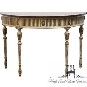 May include: A half-moon console table with a light-colored top and a dark brown trim. The table features ornate, fluted detailing and decorative carvings on the legs and apron. The table is approximately 60 inches wide.