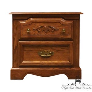 May include: A two-drawer wooden nightstand with a warm brown finish. The top drawer features a carved floral design and brass knobs. The bottom drawer has a brass handle. The nightstand has a curved base and is suitable for a bedroom.