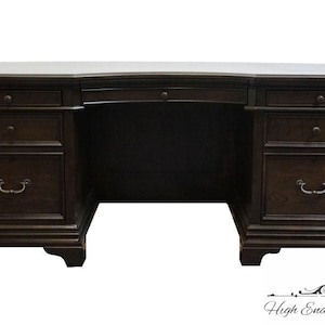 May include: Dark brown wooden desk with multiple drawers and a curved front. The desk has a classic design with decorative hardware and a large work surface. The desk is suitable for a home office or study.