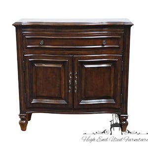 May include: A dark brown wooden cabinet featuring a single horizontal drawer at the top with two small round knobs and two lower cabinet doors with raised panel detailing and ornate silver-toned metal handles, supported by four turned feet.
