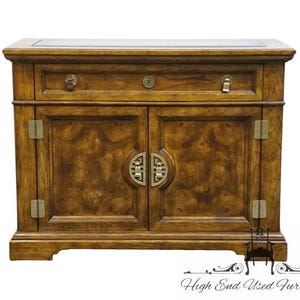 May include: A wooden cabinet with a single drawer and two doors. The cabinet is a warm brown color with a patterned wood grain. It features brass hardware, including drawer pulls and door handles. The cabinet is a piece of furniture.