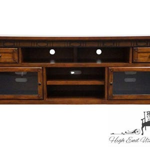 May include: A rectangular wooden media console with a medium brown finish. It features two top drawers with dark knobs, two side cabinets with glass panels, and an open center section with two shelves. The top edge has a decorative carved border.