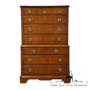 May include: A tall, wooden chest of drawers with a warm, medium-brown finish. The chest features multiple drawers with brass hardware and a classic design. The piece is likely vintage or antique, with a traditional aesthetic. The chest is suitable for bedroom storage.