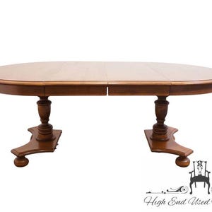May include: An oval wooden dining table with a smooth, light brown finish. The table features a central seam indicating an extendable design, supported by two sturdy pedestals, each with a turned column base and four curved feet with rounded ends.
