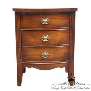 May include: A wooden nightstand with a rich brown finish, featuring three stacked drawers with curved fronts. Each drawer is equipped with a single oval brass pull handle. The piece has vertical fluted detailing on the side panels.