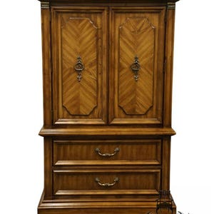 May include: A tall, wooden armoire with two doors and two drawers. The doors feature a chevron pattern and ornate handles. The armoire is a warm brown color and has decorative hardware. The bottom drawers have curved handles.