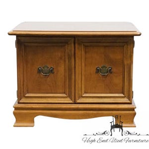 May include: A wooden cabinet with two doors and brass handles. The cabinet is a warm brown color with a light-colored top. The base has a decorative design. The cabinet is labeled "High End Used Furniture".
