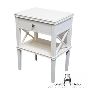 May include: A white bedside table featuring a rectangular top, a single drawer with a small round silver knob, and an open lower shelf. The sides of the table are accented with a decorative X-shaped cross brace design between the four straight legs.