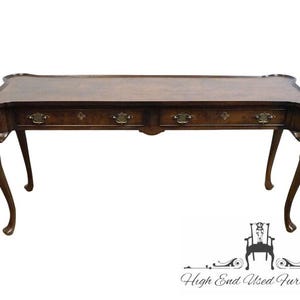 May include: A rectangular wooden desk with a dark brown finish and a smooth top surface. The desk features four drawers, each equipped with two brass-colored metal pulls. It stands on four curved legs and has a scalloped edge along the top border.