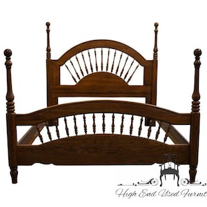 May include: A dark brown, four-poster bed frame with a headboard and footboard featuring a spindle design. The bed frame has four tall, turned posts and a curved headboard with a sunburst-like design. The bed frame is made of wood.
