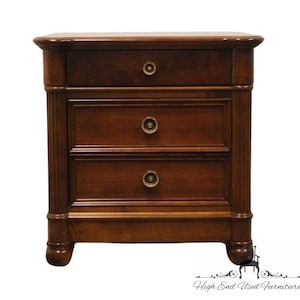 May include: A three-drawer nightstand made of dark wood. The nightstand has a rectangular shape with rounded edges and decorative brass knobs. The sides feature vertical fluted detailing. The piece is supported by short, rounded legs.