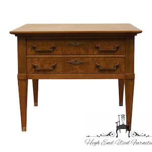 May include: A two-drawer wooden end table with a light brown finish. The table has a rectangular top and tapered legs. The drawers have brass pulls and a diamond-shaped inlay on the front.