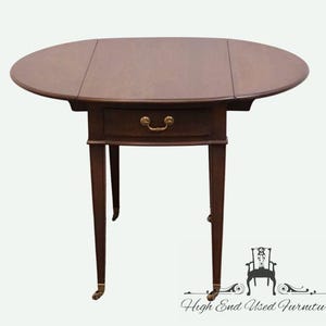 May include: A dark brown, oval drop-leaf table with a single drawer and brass hardware. The table has four slender legs with brass accents on the feet. The table is a classic piece of furniture.