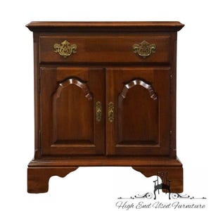 May include: A dark brown wooden nightstand with a drawer and two doors. The drawer has two ornate brass handles. The doors have decorative arched panels and brass hardware. The nightstand has a decorative base and is labeled "High End Used Furniture".