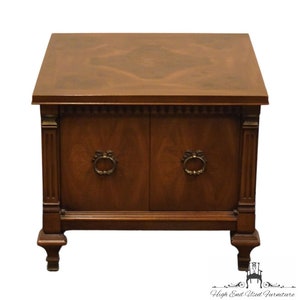 May include: A dark wood end table with two doors and decorative brass hardware. The table has a square top with a wood grain pattern and sits on four legs.