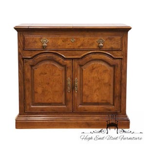 May include: A wooden cabinet with a drawer and two doors. The cabinet is made of a medium-toned wood and features ornate brass hardware. The drawer has two decorative pulls, and the doors have arched panels. The cabinet is a piece of furniture.