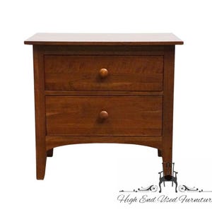 May include: A wooden nightstand with a medium brown finish, featuring a rectangular top and two stacked drawers. Each drawer is equipped with a single round wooden knob centered on the front panel. The base has an arched apron design.