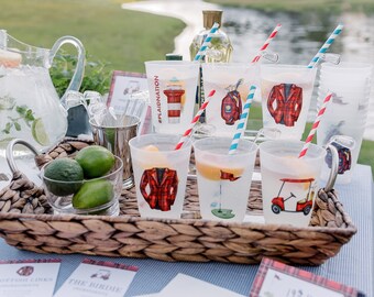 May include: A wicker tray with six clear plastic cups with red and white plaid designs. The cups have different designs, including a lighthouse, a golf bag, a golf cart, and a plaid jacket. The tray is on a blue and white checkered tablecloth. There are also three cards on the table with the text "19th Hole", "The Birdie", and "Otter Links".