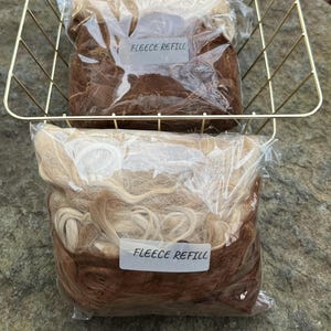 May include: A gold wire basket holds three clear plastic bags of fleece refills. The fleece is a mix of light and dark brown, with the text "FLEECE REFILL" on each label. The basket sits on a stone surface.