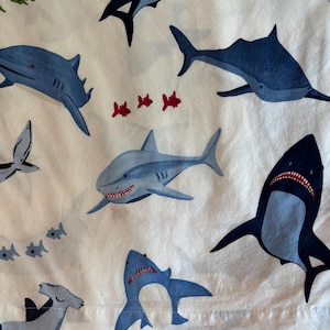 May include: White fabric with a repeating pattern of blue sharks in various poses. Some sharks have open mouths with red teeth. Small red and green fish are scattered throughout the design.