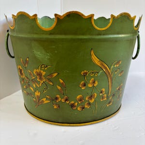 May include: A green metal planter with a scalloped rim and two handles. The planter is decorated with yellow floral and hummingbird designs. The rim and base have a gold trim.