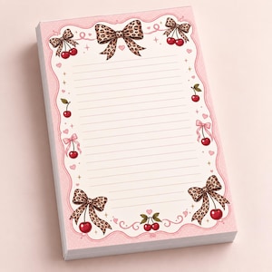 May include: A rectangular notepad with lined paper featuring a pink border decorated with red cherries, small pink hearts, and leopard print bows. The design includes scattered sparkles and a wavy frame pattern surrounding the central writing area.