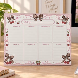 Puede incluir: Un bloc de notas semanal rectangular con borde rosa claro, decorado con lazos de estampado de leopardo e ilustraciones de cerezas rojas. El diseño incluye secciones de lunes a domingo y una columna de notas, con el texto Week of arriba.