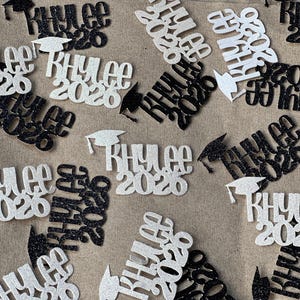 May include: Scattered graduation confetti with the name "Khylee" and the year "2020" in black and silver glitter. Some pieces include a graduation cap. The confetti is scattered on a neutral surface.