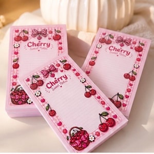 Könnte beinhalten: Drei rechteckige Papierblöcke mit einem hellrosa karierten Rand. Jeder Block trägt oben den Text Cherry Sweet Notes und ist mit Illustrationen von roten Kirschen, kleinen weißen Blumen, rosa Schleifen und einem Korb voller Kirschen verziert.