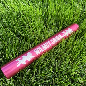May include: A magenta-colored baton lies on a bed of green grass. The baton has white text that reads "WILMOT UNION" and a logo. The baton is cylindrical and appears to be made of metal.