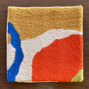 May include: A square-shaped, hand-woven rug with abstract shapes in yellow, orange, white, blue, and green.