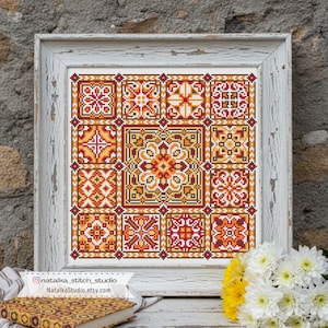May include: A framed cross-stitch artwork featuring a geometric pattern in shades of red, orange, and yellow. The design is composed of multiple square tiles with intricate floral and geometric motifs. The frame is white with a distressed finish.