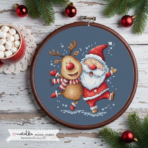 Könnte beinhalten: Ein Kreuzstich-Stickrahmen mit dem Weihnachtsmann und einem Rentier, die tanzen. Beide Figuren tragen rot-weiße Outfits vor blauem Hintergrund. Der Holzrahmen ist von Weihnachtsdekorationen umgeben, darunter Kugeln und Tannenzweige.