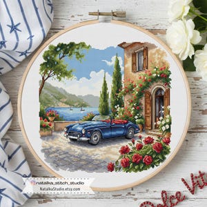 Puede incluir: Bordado de punto de cruz de un descapotable azul estacionado frente a una villa con rosas rojas. La escena incluye un lago, montañas y cipreses, todo enmarcado en un aro de madera. El texto "NataliaStudio.etsy.com" es visible.
