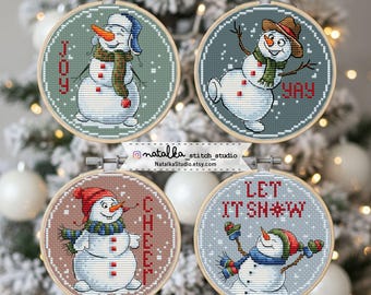 May include: Four cross-stitch embroidery hoops featuring snowmen with winter accessories. Each snowman has a unique design, with text like "JOY", "YAY", "CHEER", and "LET IT SNOW". The hoops are displayed against a blurred Christmas tree background.