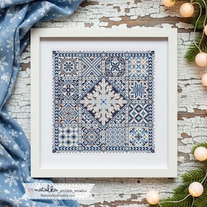 Op de afbeelding: Een ingelijst kruissteekkunstwerk met een geometrisch sneeuwvlokontwerp in blauw, wit en bruin. Het kunstwerk is in een witte lijst en staat op een verweerde houten ondergrond. Een blauwe stof met sneeuwvlokpatronen omringt het kunstwerk.