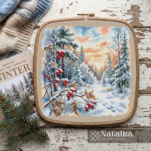 May include: A square wooden embroidery hoop containing a cross-stitch pattern of a snowy winter forest scene at sunset. The design features snow-covered pine trees and branches with bright red berries. The fabric is a light beige colour.