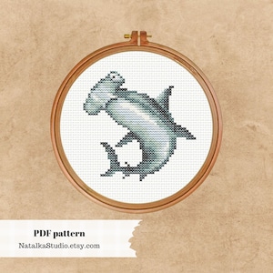 May include: A cross-stitch pattern of a hammerhead shark in a wooden embroidery hoop. The shark is gray and white and is depicted in a swimming pose. The pattern is available as a PDF download.