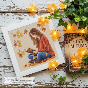 Könnte beinhalten: Ein gerahmtes Kreuzstich-Kunstwerk zeigt eine Person, die ein Buch liest, umgeben von Herbstblättern. Die Person trägt einen orangefarbenen Pullover und blaue Jeans. Ein Buch mit dem Titel "Cozy Autumn" und Lichterketten in Blattform sind ebenfalls zu sehen.