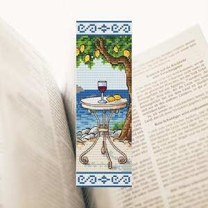 Pode incluir: Um marcador de página de ponto cruz que representa uma mesa com uma taça de vinho tinto e limões sobre uma toalha de mesa branca. A mesa está situada em frente a um fundo de mar azul e uma árvore de limão com folhas verdes e limões amarelos.
