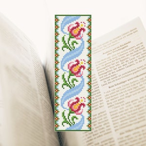 May include: A cross-stitch bookmark with a white background and a blue, green, and pink floral design. The bookmark features three pink flowers with yellow centers and green leaves. The flowers are arranged in a vertical pattern, with the stems winding around each other.